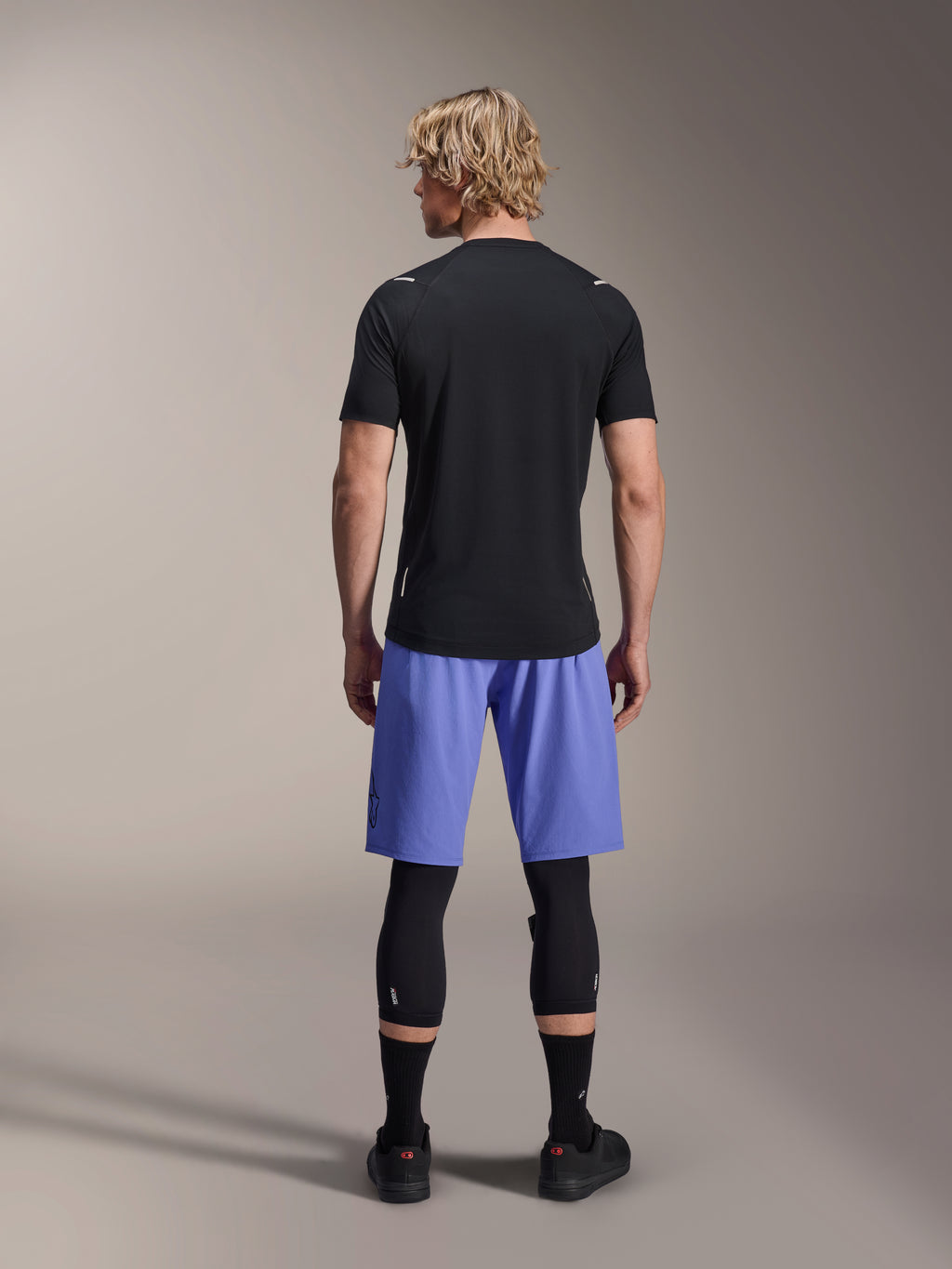 Mannelijke rijder in Alpinestars A-Aria Lite Shorts, MTB-short, Baja Blue, achteraanzicht, gecombineerd met een zwarte jersey met korte mouwen, zwarte kniebeschermers en zwarte fietsschoenen, hoogwaardige mountainbikekleding
