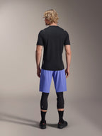 Mannelijke rijder in Alpinestars A-Aria Lite Shorts, MTB-short, Baja Blue, achteraanzicht, gecombineerd met een zwarte jersey met korte mouwen, zwarte kniebeschermers en zwarte fietsschoenen, hoogwaardige mountainbikekleding