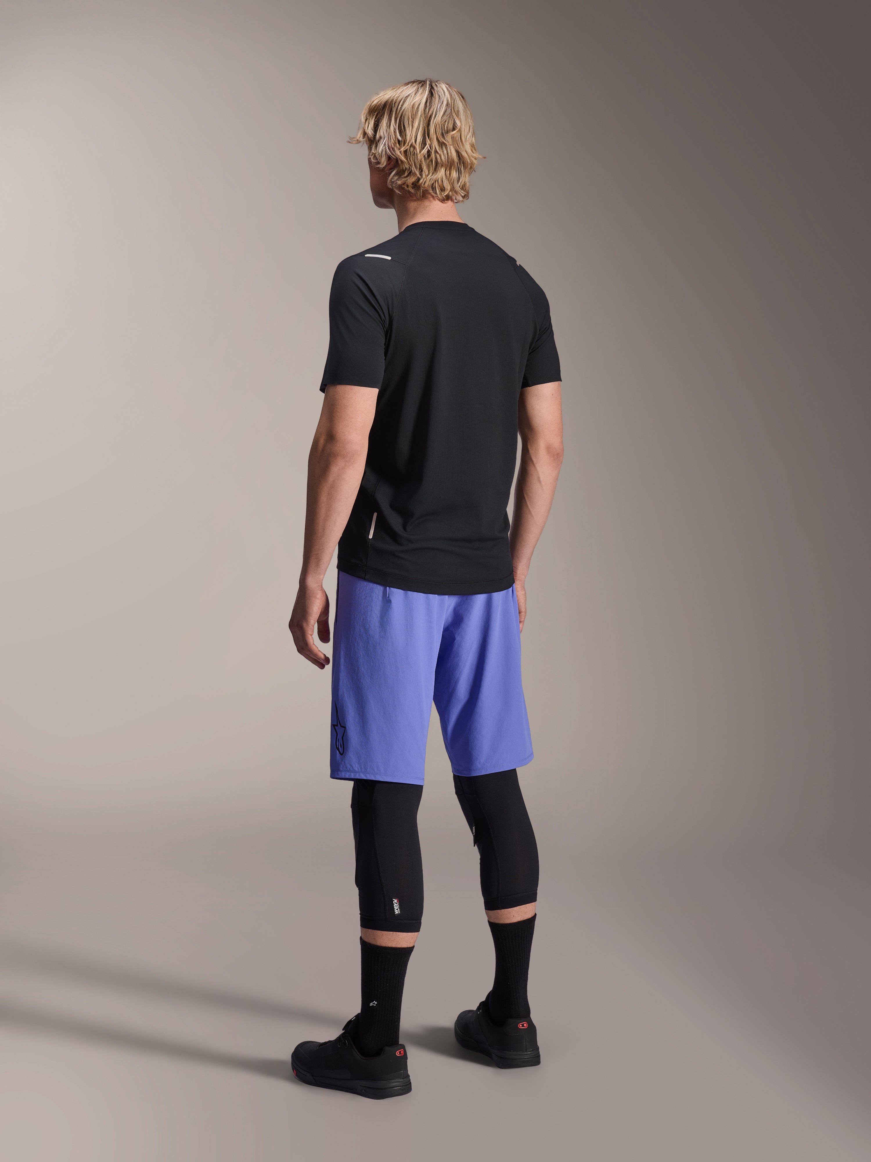 Mannelijk model van achteren in Alpinestars A-Aria Lite Shorts in Baja Blue, gecombineerd met een zwarte jersey met korte mouwen en zwarte kniebeschermers, professionele MTB-fietskleding voor trailrijden.