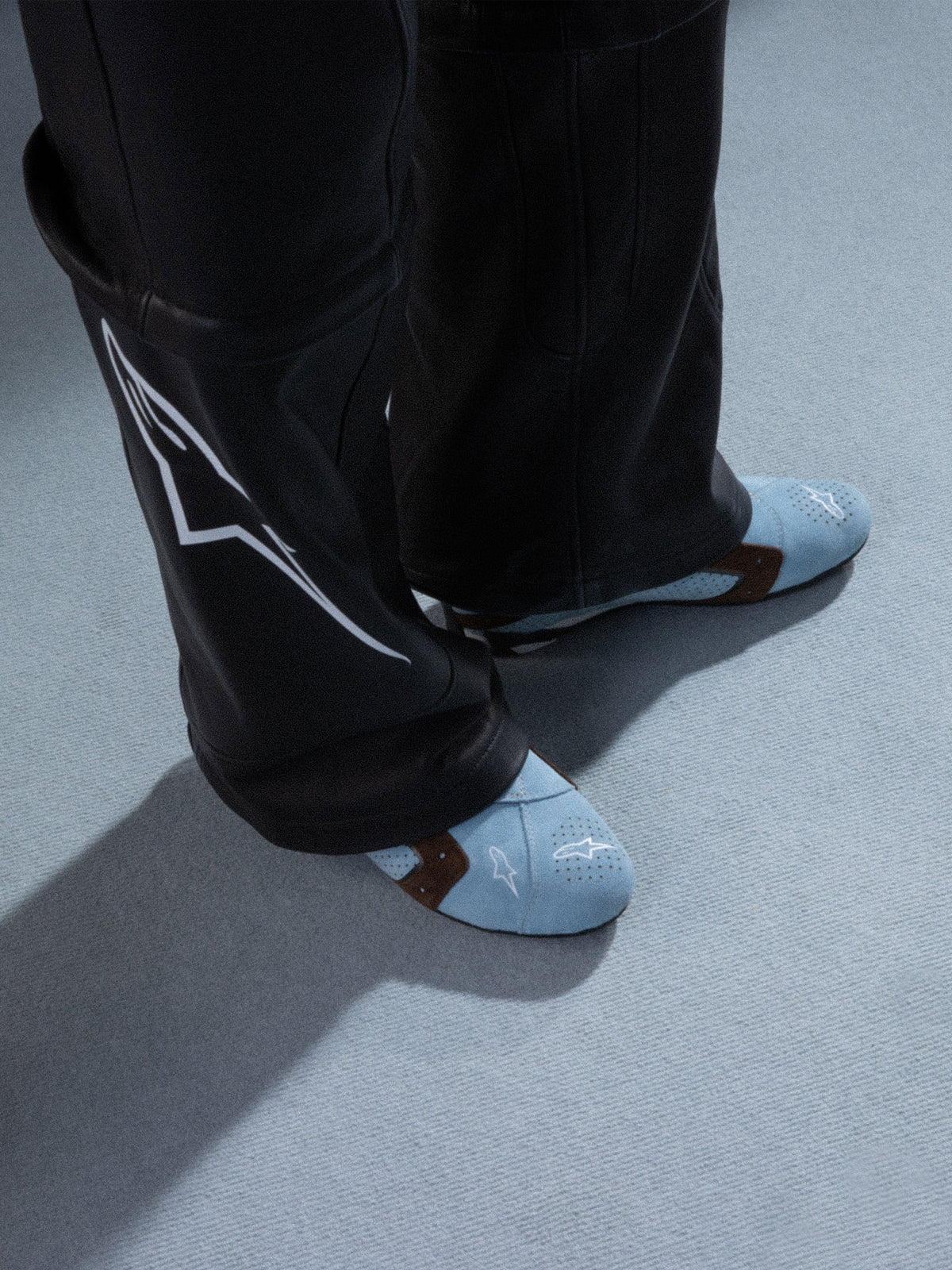 Alpinestars RSRV Starline schoenen, lichtblauwe sneakers met geperforeerde patronen, bruine accenten, wit logo, premium lederen constructie, gedragen met zwarte leren broek met wit ontwerp