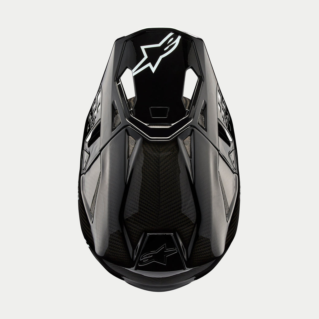 Alpinestars Supertech M10 Solid helm, motorcrosshelm, zwart glanzend carbon, bovenaanzicht met zichtbaar koolstofvezelweefsel, wit Astars-logo op de klep, slank aerodynamisch profiel met geïntegreerde ventilatieopeningen