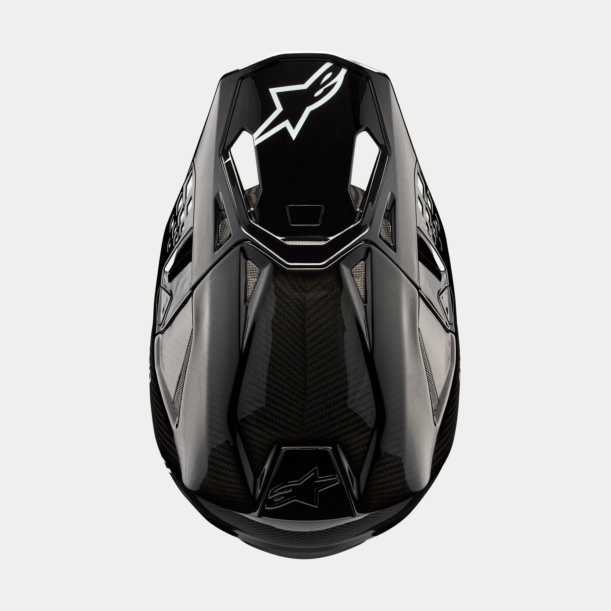 Alpinestars Supertech M10 Solid helm, motorcrosshelm, zwart glanzend carbon, bovenaanzicht met zichtbaar koolstofvezelweefsel, wit Astars-logo op de klep, slank aerodynamisch profiel met geïntegreerde ventilatieopeningen