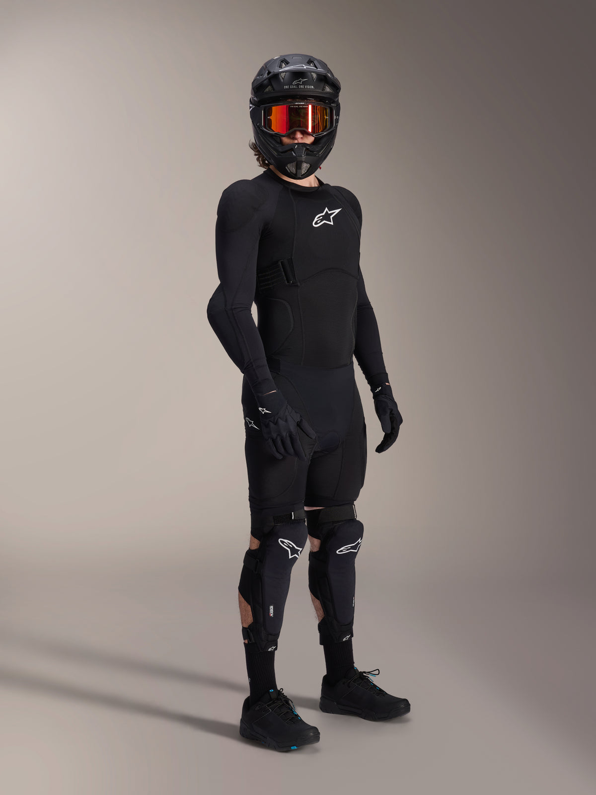 Alpinestars A-Impact Plasma Pro knie- en scheenbeschermers, zwart en wit, gedragen door een rijder met een volledige mountainbike-beschermingsoutfit, inclusief een zwarte gepantserde top met lange mouwen, zwarte handschoenen en een full-face helm met oranje bril.