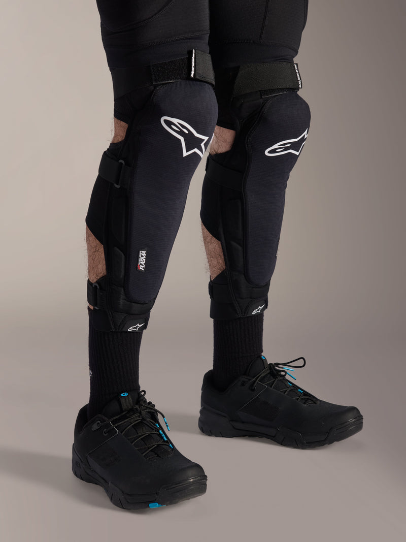 Alpinestars A-Impact Plasma Pro kniebeschermers, zwart en wit, met Nucleon PLASMA-technologie branding, gedragen met zwarte sokken en zwarte mountainbikeschoenen, stevige klittenbandsluitingen, ergonomische beschermingsuitrusting voor off-road fietsen