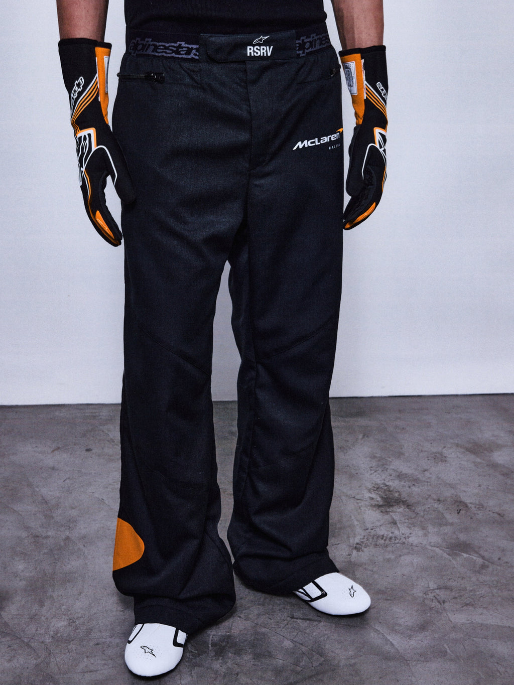 Alpinestars RSRV Arena Suit racingbroek, zwart met oranje details, McLaren-logo's, premium motorsport pantalon, gedragen met race-uitrusting, getextureerde grijze ondergrond
