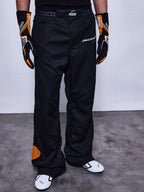 Alpinestars RSRV Arena Suit racingbroek, zwart met oranje details, McLaren-logo's, premium motorsport pantalon, gedragen met race-uitrusting, getextureerde grijze ondergrond