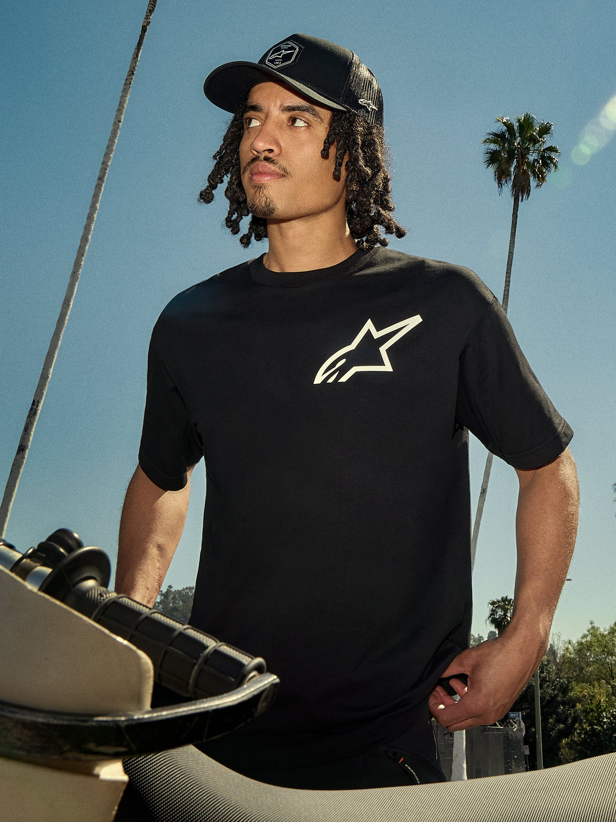 Alpinestars One Oversized CSF Tee, T-shirt met korte mouwen, zwart met wit Astars-logo, gedragen door een man met een zwarte truckerpet, outdoor lifestyle setting met palmbomen en een motorfiets, casual motorsport streetwear