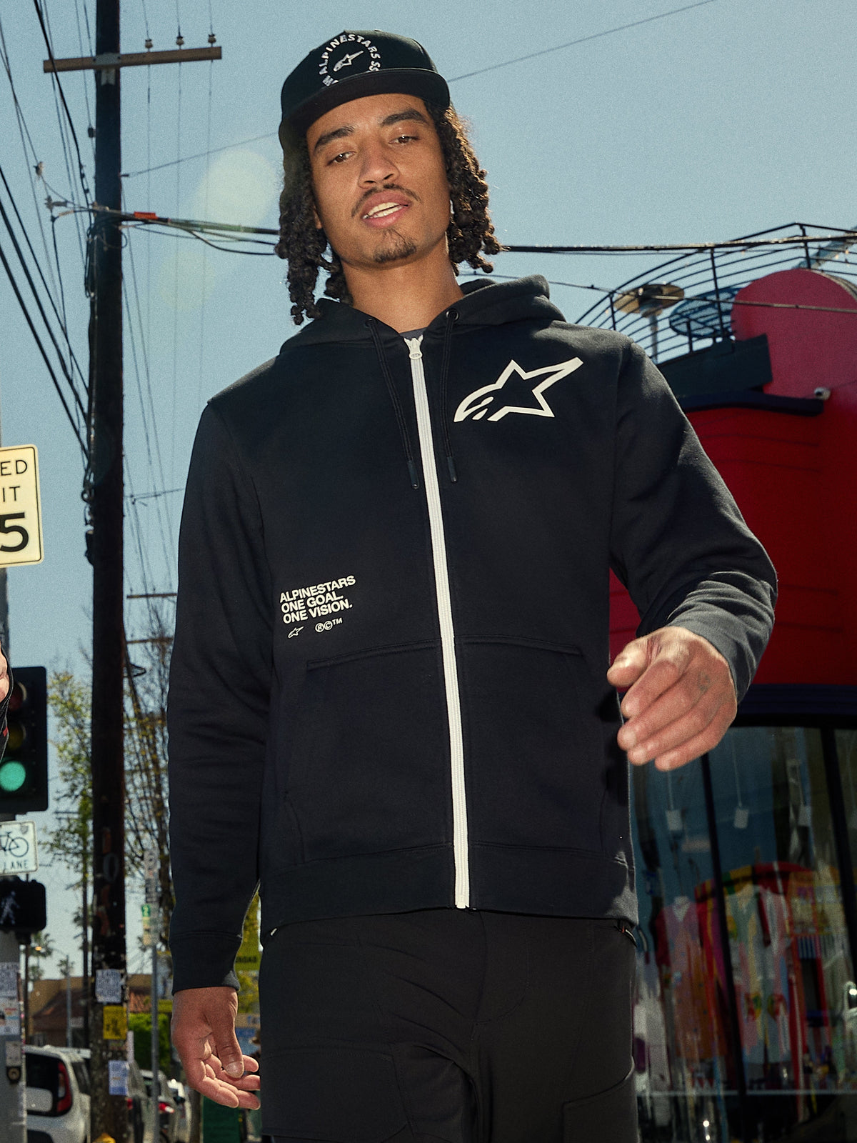 Alpinestars Transverse Hoodie, zwarte fleece met rits aan de voorzijde, witte rits en wit Astars-logo op de borst, 'One Goal One Vision' tekstprint, casual streetwear-stijl, gedragen door een man in een stedelijke buitenomgeving