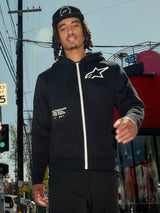 Alpinestars Transverse Hoodie, zwarte fleece met rits aan de voorzijde, witte rits en wit Astars-logo op de borst, 'One Goal One Vision' tekstprint, casual streetwear-stijl, gedragen door een man in een stedelijke buitenomgeving