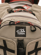 Alpinestars AT-15 Rugzak, Aluminum Walnut, close-up van de All Terra logo-patch op beige technisch textiel, voorzien van oranje trekkoorden aan de ritsen en robuuste constructiedetails