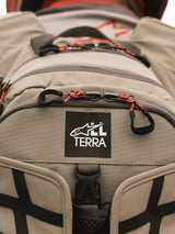 Alpinestars AT-15 Rugzak, Aluminum Walnut, close-up van de All Terra logo-patch op beige technisch textiel, voorzien van oranje trekkoorden aan de ritsen en robuuste constructiedetails