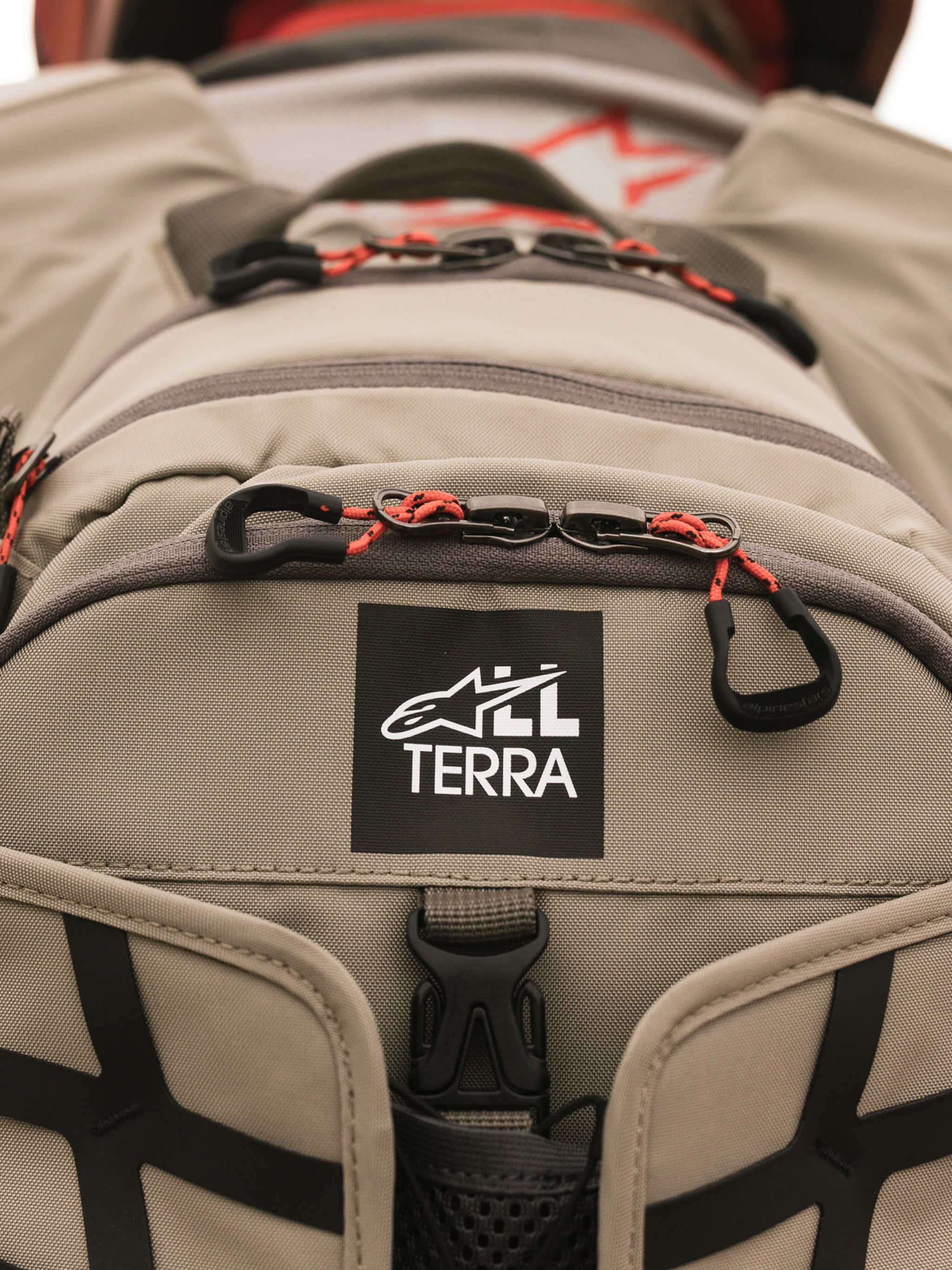 Alpinestars AT-15 Rugzak, Aluminum Walnut, close-up van de All Terra logo-patch op beige technisch textiel, voorzien van oranje trekkoorden aan de ritsen en robuuste constructiedetails