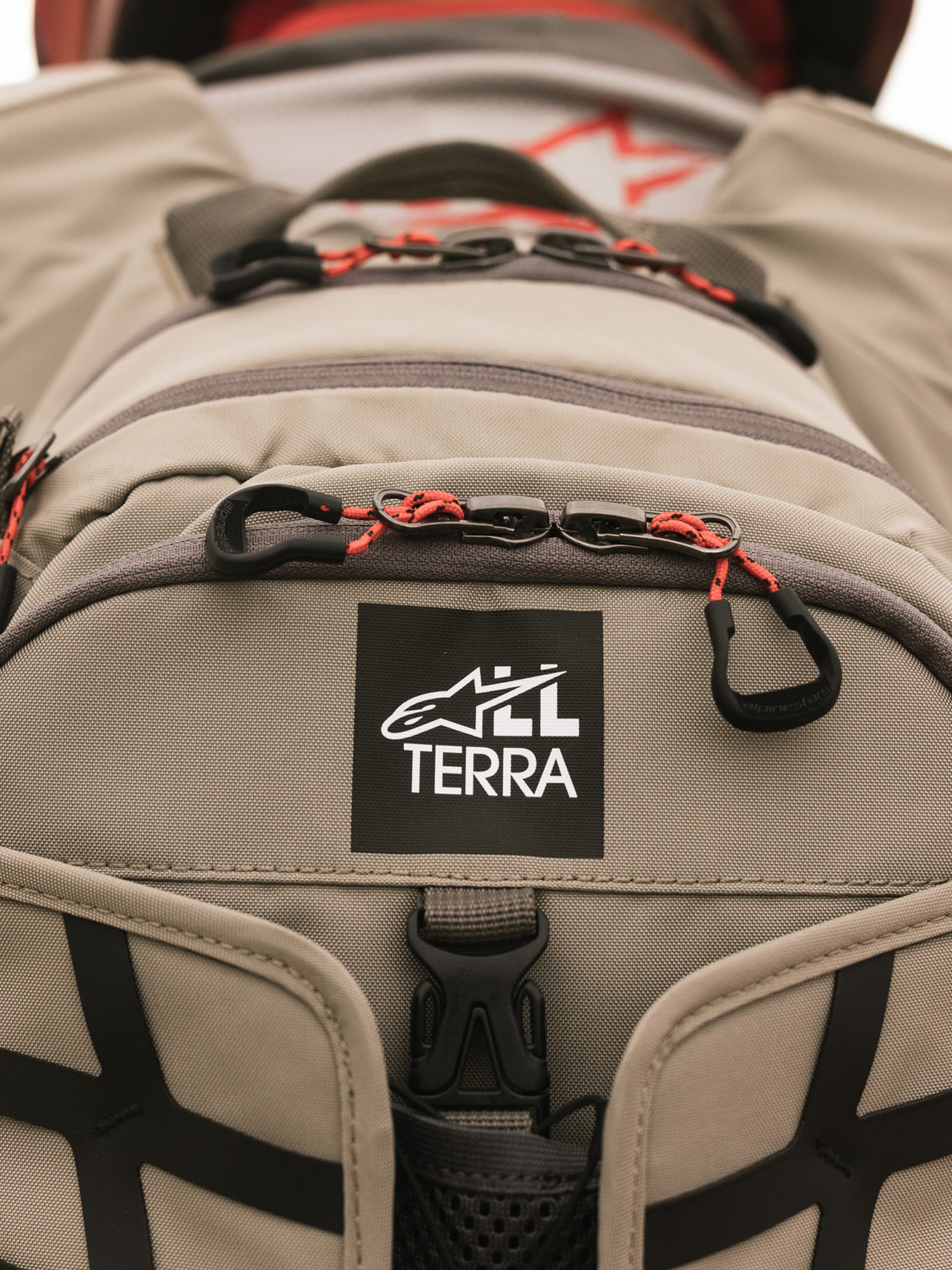 Alpinestars AT-15 Rugzak, Aluminum Walnut, close-up van de All Terra logo-patch op beige technisch textiel, voorzien van oranje trekkoorden aan de ritsen en robuuste constructiedetails