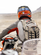 Alpinestars AT-15 rugzak in Aluminum Walnut, beige technische motorrugzak met zwarte banden en oranje ritsaccenten, 'All Terra' branding, gedragen door een adventure motorrijder in een woestijnlandschap met een rood-oranje helm en grijze motoruitrusting.