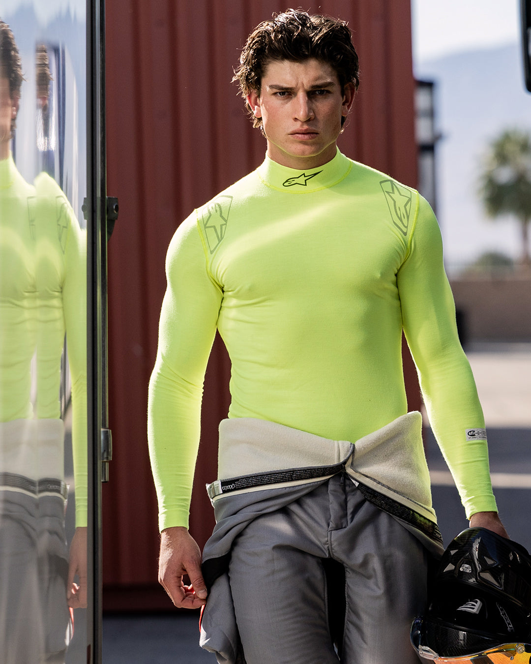 Alpinestars ZX Evo V3 Top - Lange Mouwen, auto racing base layer, Yellow Fluo, gedragen door een mannelijke coureur met een grijs racepak om zijn middel geknoopt, houdt een zwarte helm vast, voorzien van Alpinestars-logo's op de nek en schouders, technische vochtafvoerende kleding voor de autosport