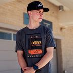 Alpinestars Galun CSF Tee, T-shirt met korte mouwen, antraciet, vintage benzineblik-graphic aan de voorkant met 'astars' branding en 'MCMLXIII' Romeinse cijfers, gedragen door een man met een zwarte pet met logo, casual motorsport lifestyle
