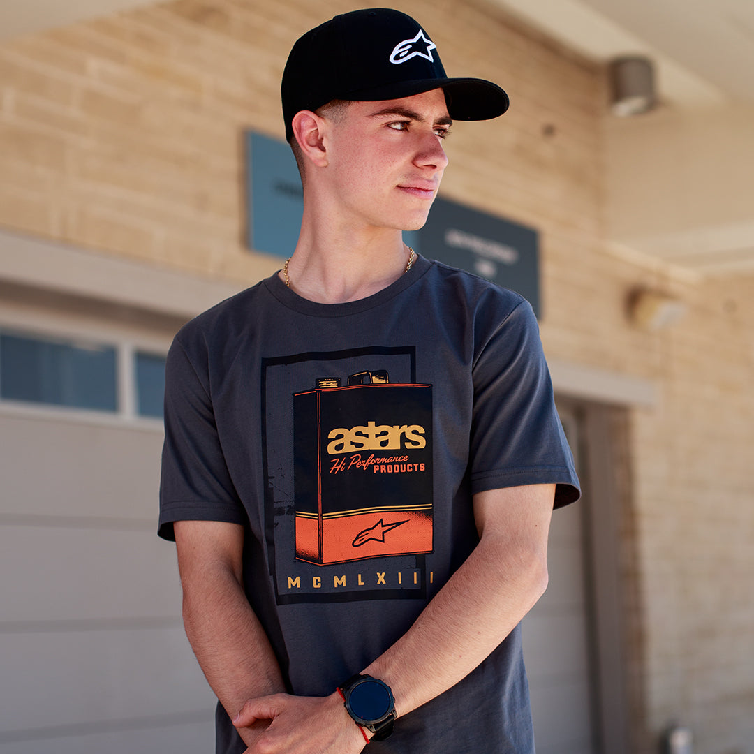 Alpinestars Galun CSF Tee, T-shirt met korte mouwen, antraciet, vintage benzineblik-graphic aan de voorkant met 'astars' branding en 'MCMLXIII' Romeinse cijfers, gedragen door een man met een zwarte pet met logo, casual motorsport lifestyle