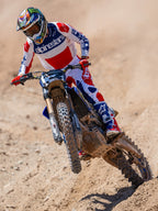 Motorcrossrijder in actie op een crossbaan, draagt de Limited Edition Techstar Fortitude Jersey in helderrood, wit en blauw, gecombineerd met bijpassende crossbroek en laarzen, opvallende Alpinestars-branding, professionele off-road racekleding
