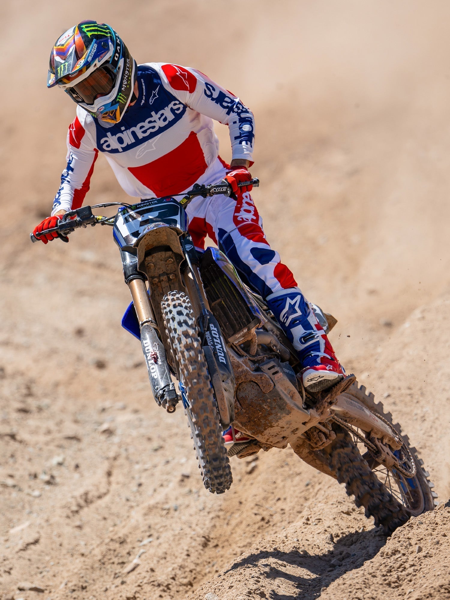 Alpinestars Limited Edition Techstar Fortitude crossbroek, motorcross broek, felrood, wit en blauw, gedragen door een rijder in actie op een zandcircuit, inclusief bijpassende jersey en laarzen, prestatiegerichte off-road race-uitrusting.