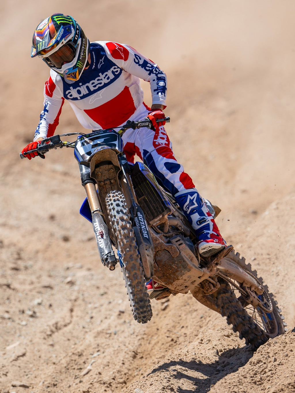 Motorcrossrijder in actie met Alpinestars Limited Edition Fortitude Tech 10 laarzen, helderrood, wit en blauw race-shirt en -broek, gecoördineerde patriottische kledingset voor off-road prestaties op een dirt track.