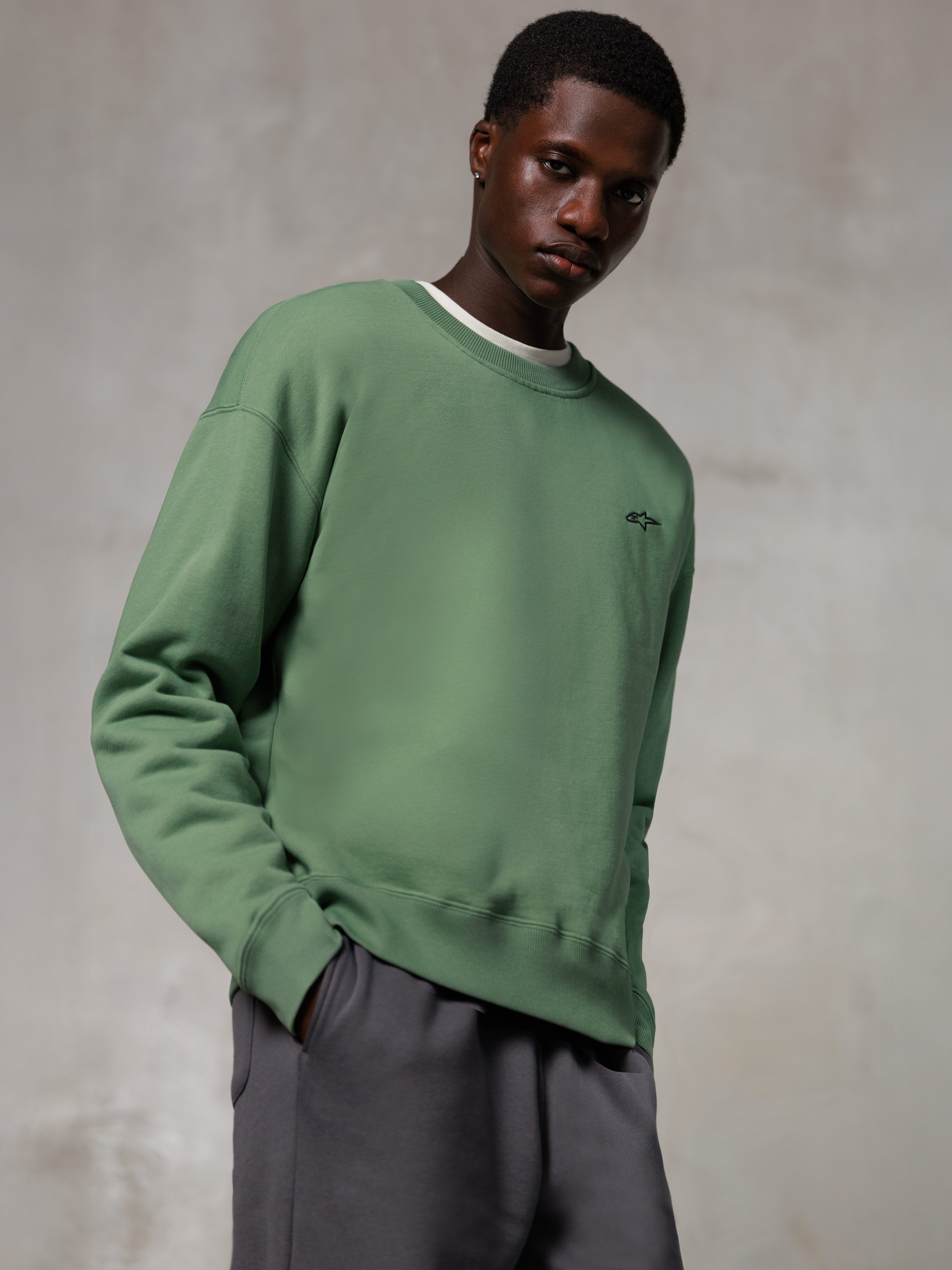 Alpinestars Essentials Ageless Crew, sweatshirt met ronde hals, cactusgroen, met een klein zwart Alpinestars-logo op de borst, gedragen door een model met een grijze broek, casual lifestyle streetwear