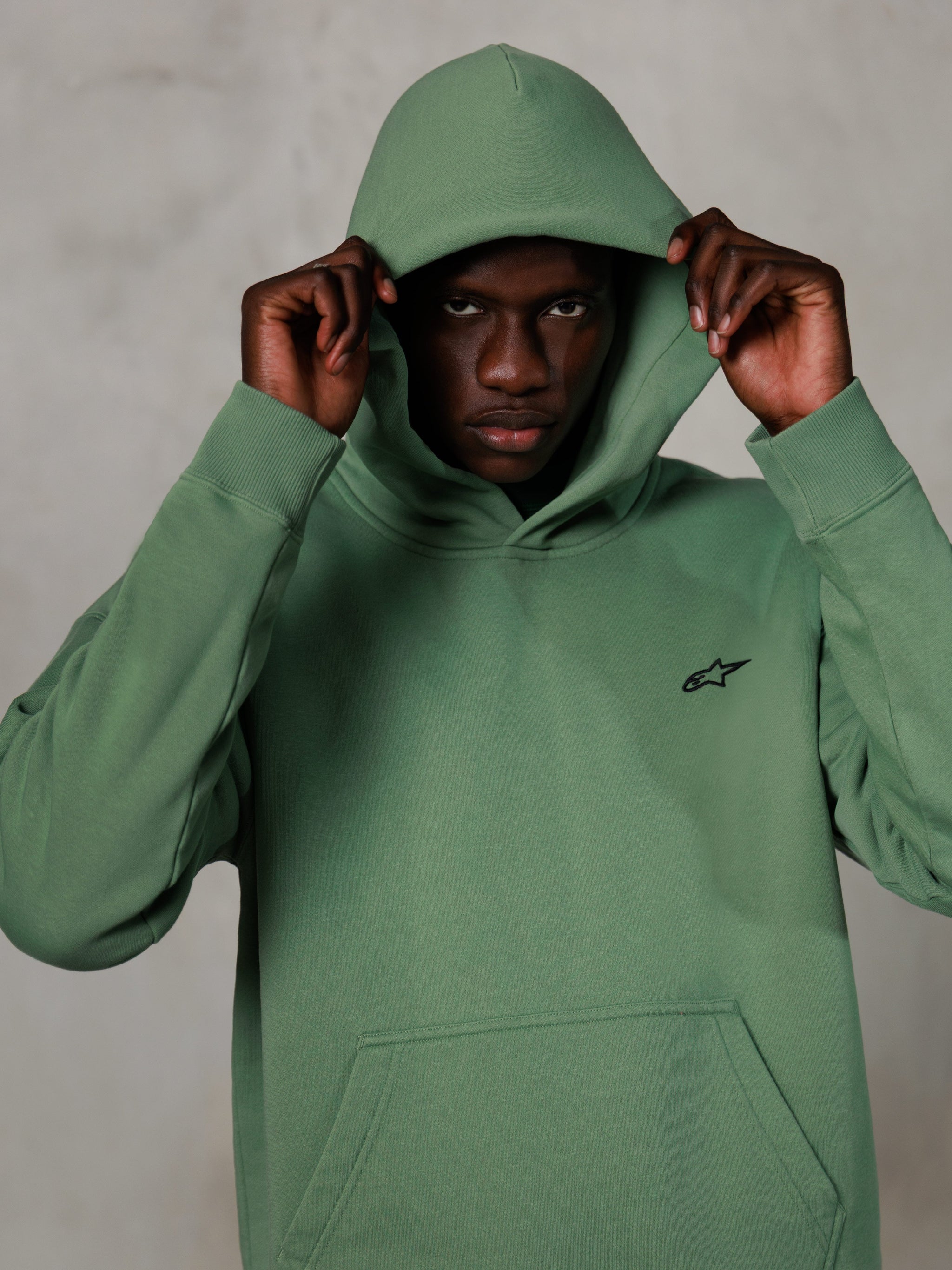 Alpinestars Essentials Ageless Hoodie, casual streetwear, cactusgroen met klein zwart logo op de borst, model draagt hoodie met de capuchon op, kangoeroezak aan de voorzijde, ontspannen op motorsport geïnspireerd ontwerp