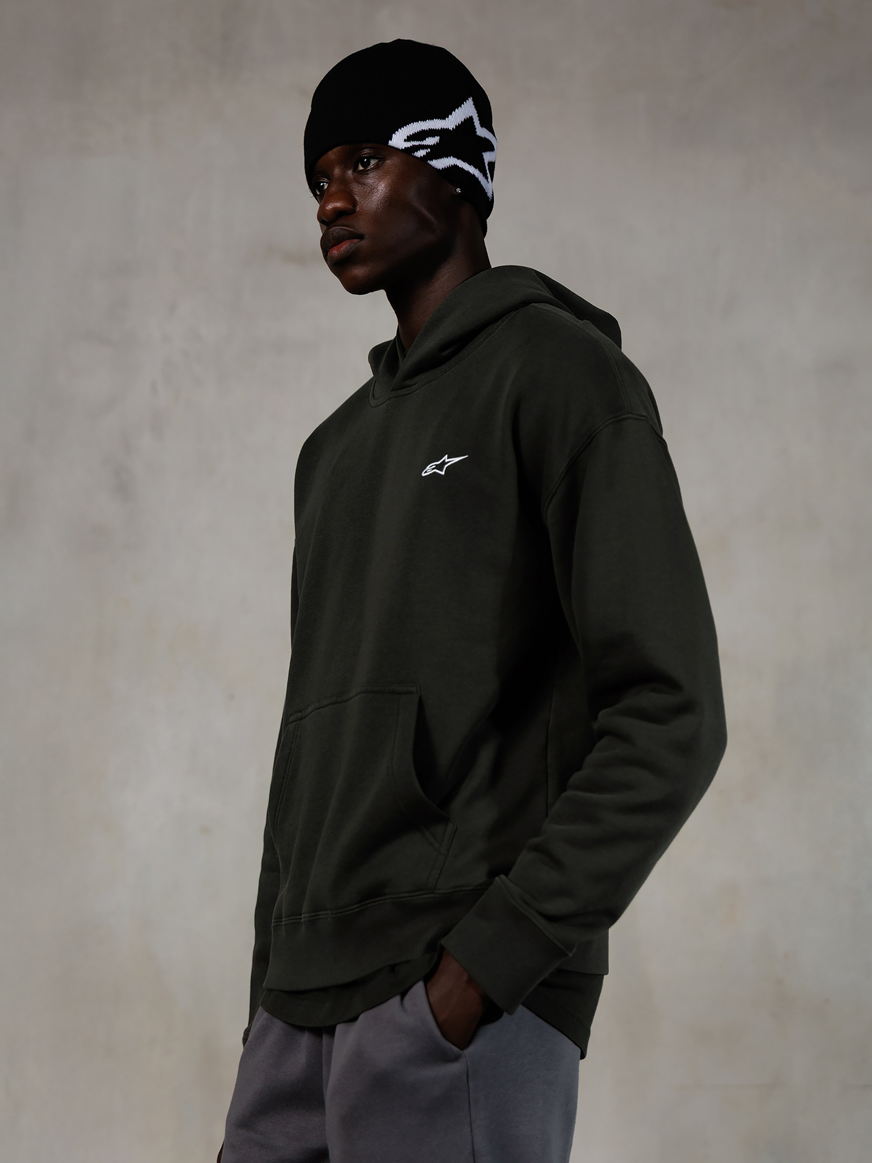 Alpinestars Essentials Ageless Hoodie, Off Black, mannelijk model in casual streetwear, donker antraciet stone-washed fleece met een klein wit ster-logo, gecombineerd met een zwarte Alpinestars beanie en een grijze broek