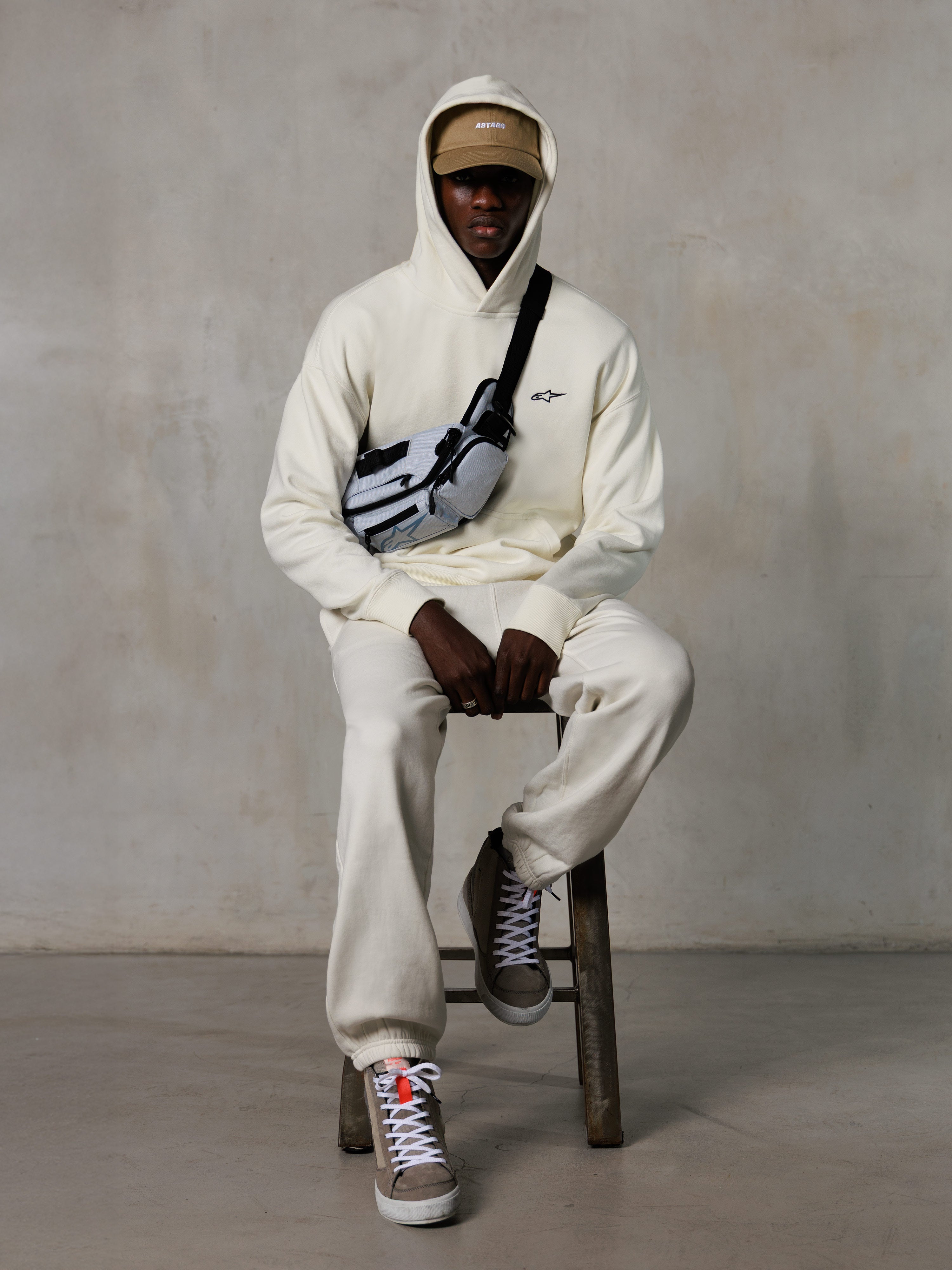 Alpinestars Essentials Ageless Hoodie, Off-White, gedragen door een model zittend op een kruk met bijpassende fleece joggingbroek, beige ASTARS-pet en crossbodytas, casual op motorsport geïnspireerde streetwear