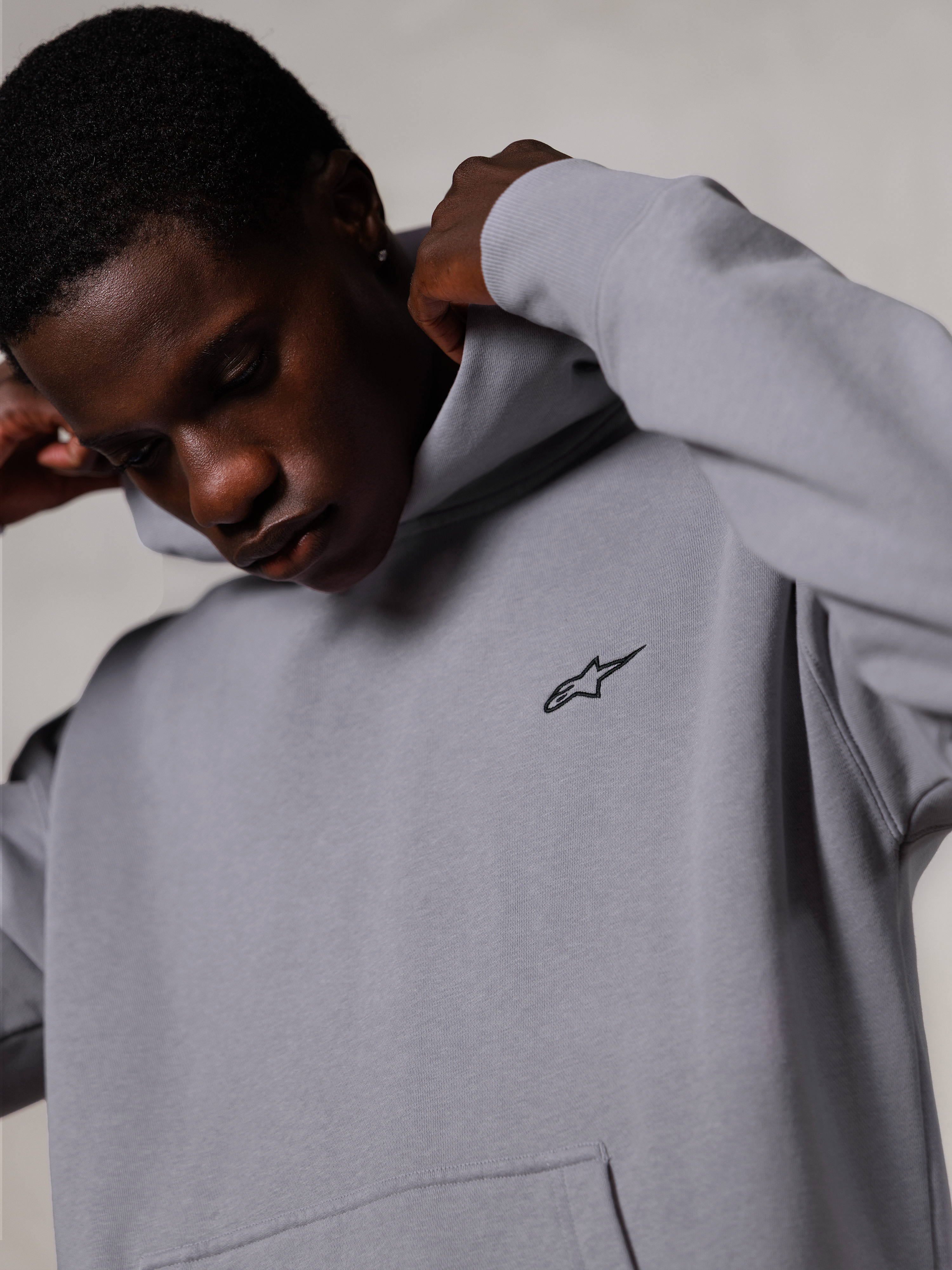 Alpinestars Essentials Ageless Hoodie, Silver, close-up van een man die de capuchon van de grijze hoodie verstelt, klein zwart Alpinestars-logo op de borst, premium op de motorsport geïnspireerde vrijetijdskleding