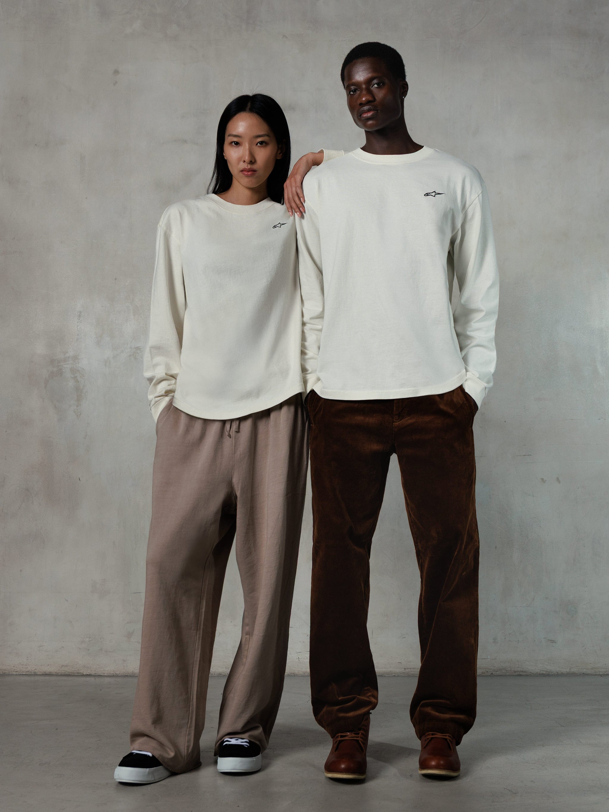 Alpinestars Essentials Ageless LS Tee, T-shirt met lange mouwen, Off White, gedragen door een mannelijk en vrouwelijk model, klein zwart Alpinestars-logo op de borst, relaxed fit, gestyled met een beige joggingbroek en bruine corduroy broek, casual op motorsport geïnspireerde streetwear