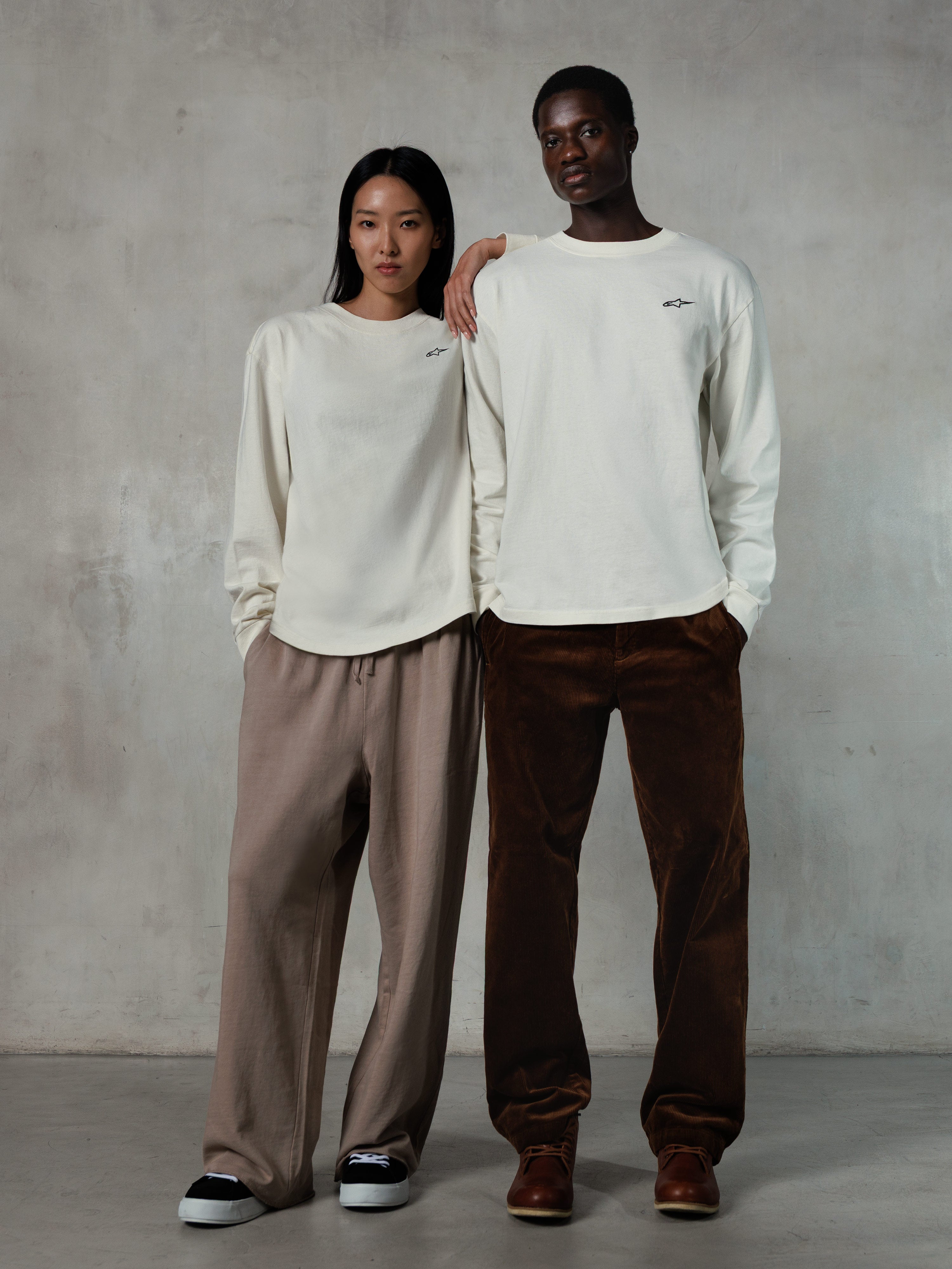 Alpinestars Essentials Ageless LS Tee, T-shirt met lange mouwen, Off White, gedragen door een mannelijk en vrouwelijk model, klein zwart Alpinestars-logo op de borst, relaxed fit, gestyled met een beige joggingbroek en bruine corduroy broek, casual op motorsport geïnspireerde streetwear