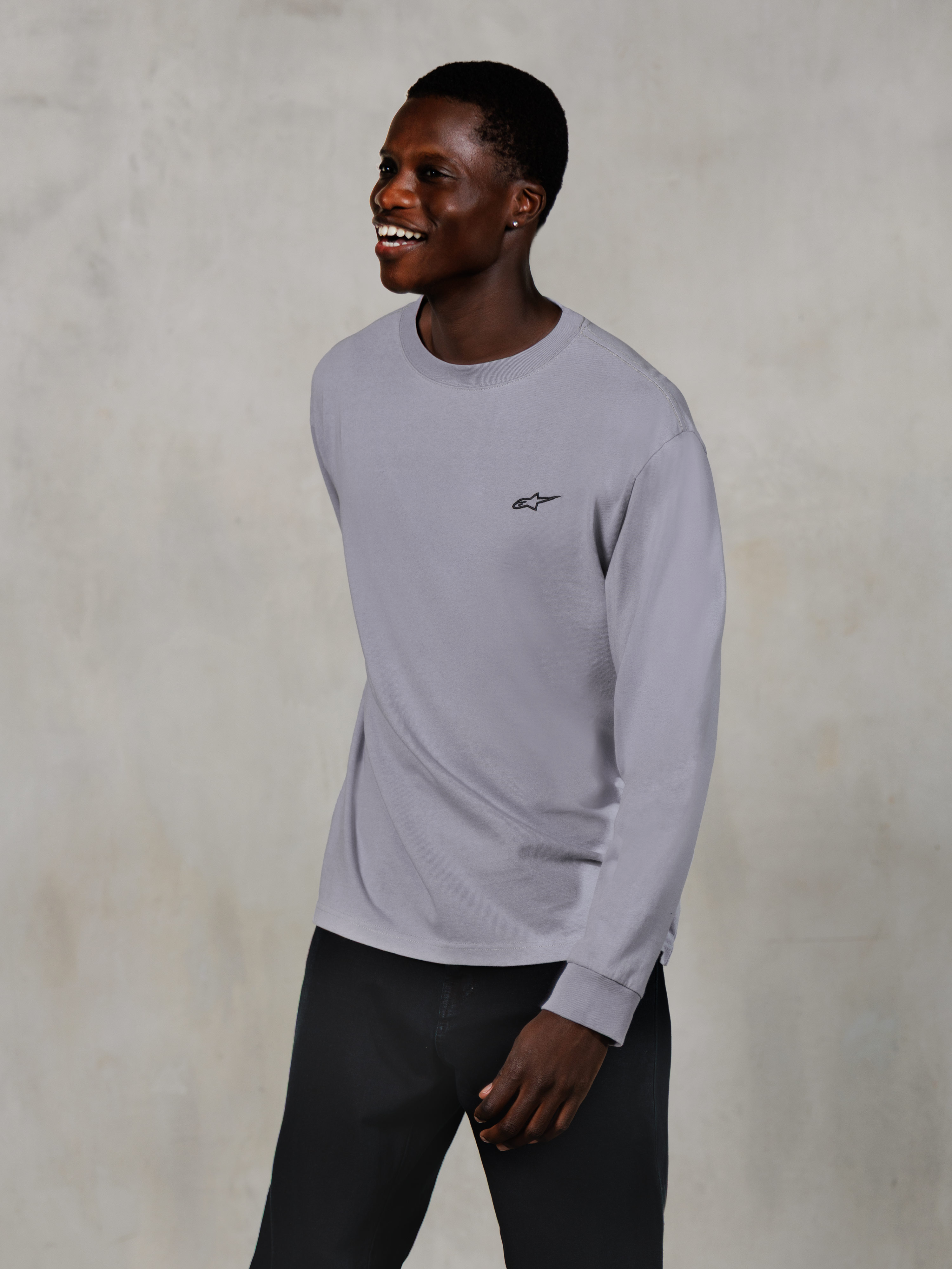 Alpinestars Essentials Ageless LS Tee, T-shirt met lange mouwen, zilver, klein zwart logo op de borst, gedragen door een glimlachend model met donkere broek, ontspannen eigentijdse pasvorm, casual motorsport lifestyle-kleding