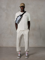 Alpinestars Essentials Ageless SS Tee, T-shirt met korte mouwen, Off White, met een klein zwart logo op de borst, gedragen met bijpassende joggingbroek, beige baseballpet en een grijze crossbody tas, casual lifestyle streetwear