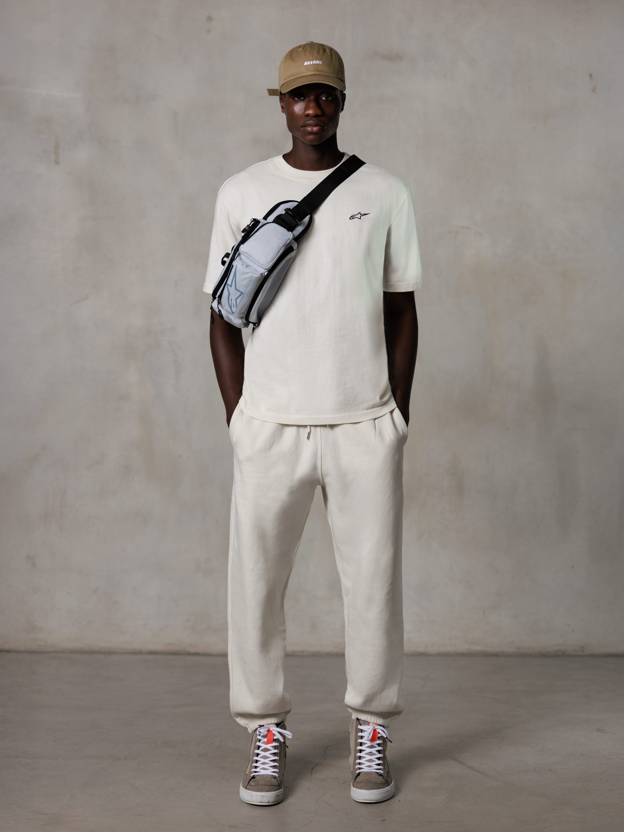 Alpinestars Essentials Ageless SS Tee, T-shirt met korte mouwen, Off White, met een klein zwart logo op de borst, gedragen met bijpassende joggingbroek, beige baseballpet en een grijze crossbody tas, casual lifestyle streetwear