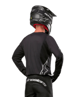 Alpinestars 2024 Fluid Graphite Gear Set, motorcross shirt en broek, grafiet en zilver, achteraanzicht met zilveren logo's op de onderrug en taille, gedragen met zwarte helm met carbon-effect, lichtgewicht poly-stof ontwerp voor off-road rijden
