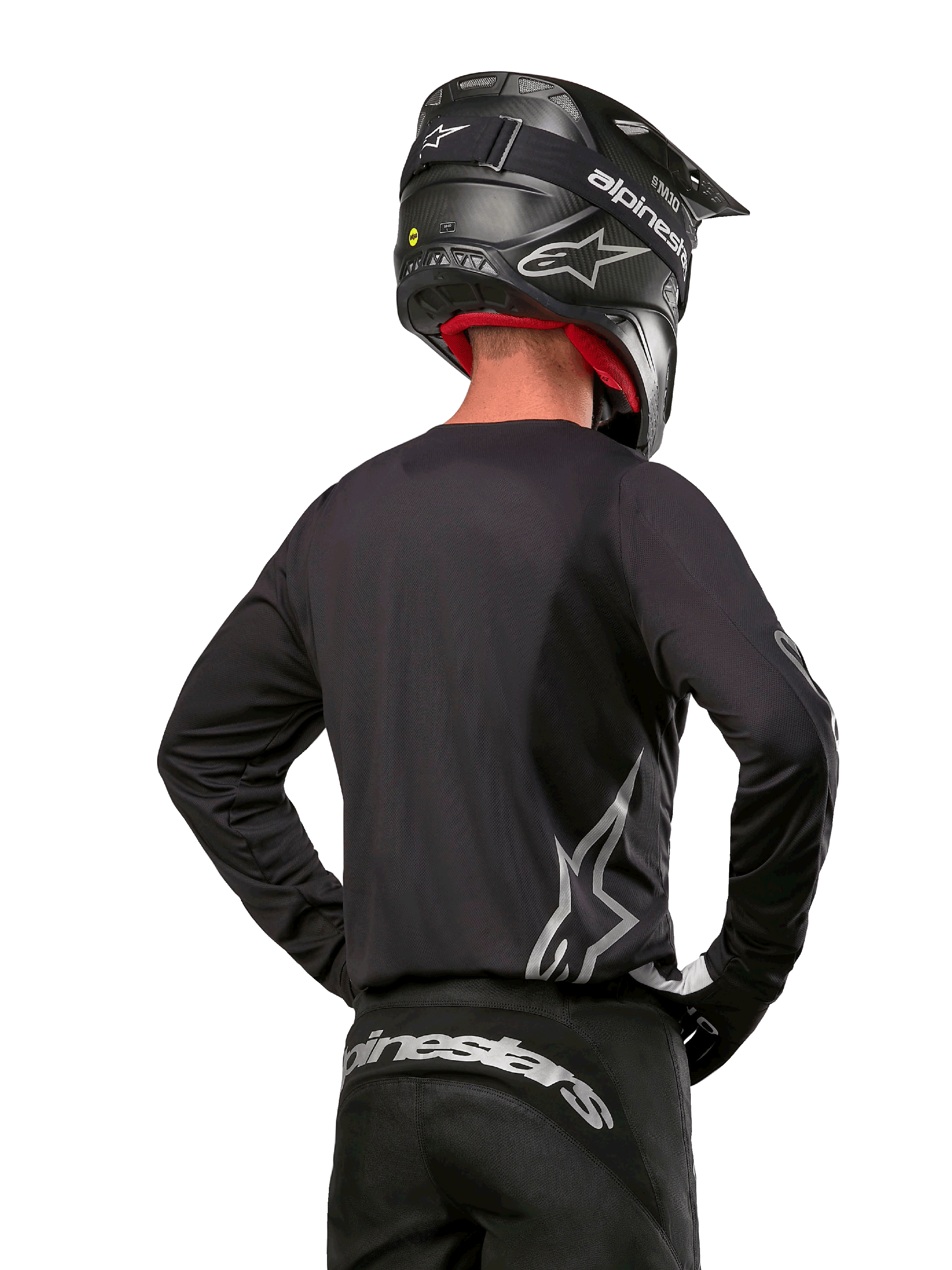 Alpinestars 2024 Fluid Graphite Jersey in Zwart Zilver, motorcrosser van achteren gezien met een groot zilveren logo op de zijkant, gecombineerd met een zwarte helm met carbon-textuur en zwarte broek, professionele off-road race-uitrusting