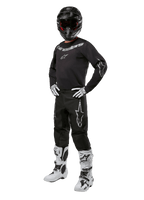 Alpinestars 2024 Fluid Graphite Gear Set, complete motorcross outfit, zwart met grijze en witte logo's, inclusief jersey en broek gedragen met bijpassende zwarte helm, witte en zwarte handschoenen en laarzen, ontworpen voor off-road bescherming