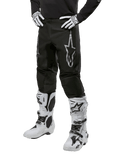 Alpinestars 2024 Fluid Graphite crossbroek, motorcross broek, zwart en zilver, met een zilveren Alpinestars-logo op het bovenbeen, getoond met witte motorcrosslaarzen en handschoenen, ontworpen voor off-road motorrijden