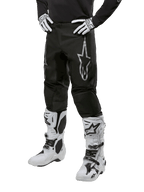 Alpinestars 2024 Fluid Graphite crossbroek, motorcross broek, zwart en zilver, met een zilveren Alpinestars-logo op het bovenbeen, getoond met witte motorcrosslaarzen en handschoenen, ontworpen voor off-road motorrijden