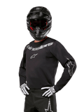 Alpinestars 2024 Fluid Graphite Jersey, zwart met zilver motorcross shirt, lange mouwen met sportieve pasvorm, gedragen met zwarte helm en witte handschoenen, voorzien van zilveren Alpinestars-branding, off-road motoruitrusting