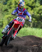 Motocross-rijder op een rode Honda crossmotor, gekleed in de Alpinestars 2025 Fluid Wurx Jersey in paars en felrood, bijpassende broek, witte laarzen en een Red Bull-helm, die een bocht neemt op een zandcircuit.