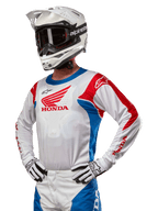 Motorcrossrijder die de Honda Racer Iconic Jersey draagt in wit, felblauw en felrood, met een groot Honda-vleugellogo op de borst, gecombineerd met een witte Alpinestars-helm en bijpassende wit-zwarte handschoenen, gecoördineerde uitrusting voor off-road prestaties