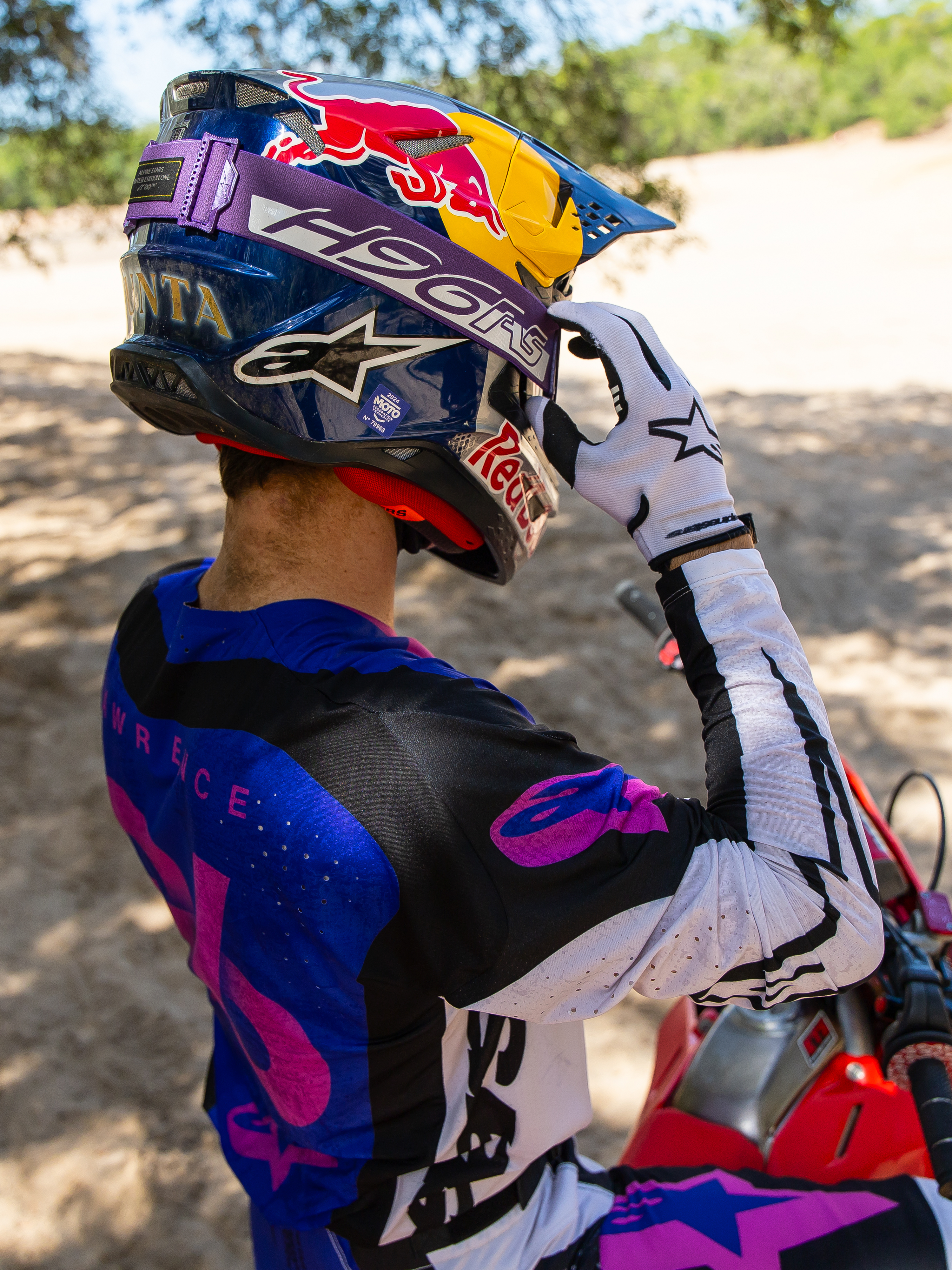 Motocrossrijder Hunter Lawrence met Limited Edition HUNTA96 XXV Supertech Vision crossbril, paarse band met 'H96' en gele stiksels, blauwe Red Bull-helm, paars-wit jersey met zwarte accenten, witte handschoenen, stelt zijn bril af in een zanderige off-road omgeving