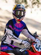 Motorcrosser op een rode crossmotor, Limited Edition HUNTA96 XXV Supertech Vision Goggles in paars en wit met zilveren spiegellenzen, bijpassende paarse, zwarte en witte Alpinestars jersey en broek, Red Bull-helm, witte handschoenen, gecoördineerde professionele motorcrossuitrusting.