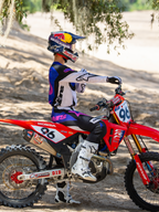 Motorcrosser met de Limited Edition HUNTA96 XXV Supertech Vision Goggles in Purple White Mirror Silver, bijpassende paars-witte motorcross jersey en witte laarzen, zittend op een rode Honda crossmotor met nummer 96, outdoor off-road setting op een zanderig circuit.