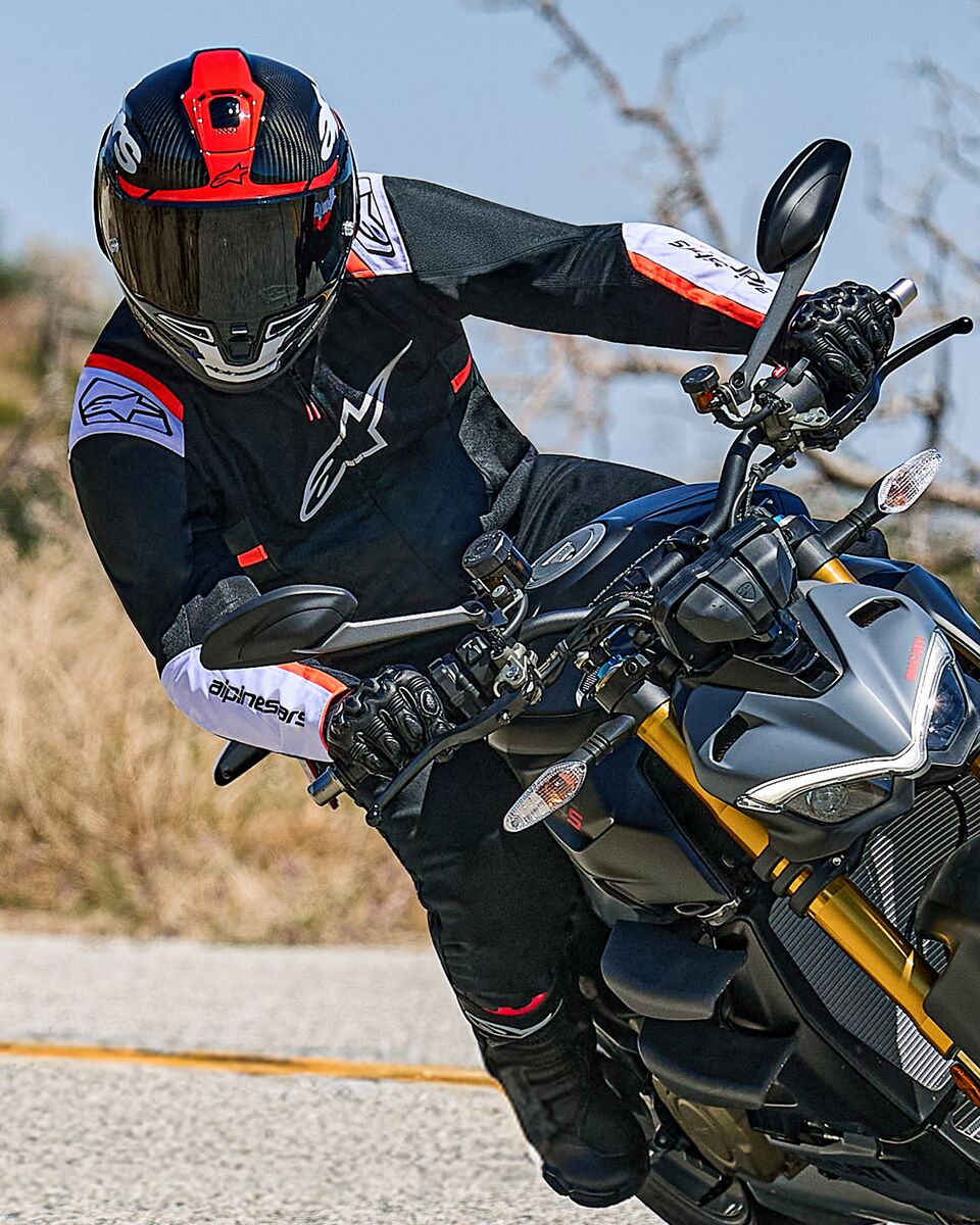 Motorrijder die in een bocht leunt op een naked sportmotor, draagt de Alpinestars T-SPS Air v2 Jas in zwart met witte en rood-fluo accenten, gecombineerd met een zwart-rode helm en zwarte handschoenen, prestatiegerichte textiele uitrusting voor stedelijk sportief rijden.