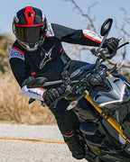 Motorrijder die in een bocht leunt op een naked sportmotor, draagt de Alpinestars T-SPS Air v2 Jas in zwart met witte en rood-fluo accenten, gecombineerd met een zwart-rode helm en zwarte handschoenen, prestatiegerichte textiele uitrusting voor stedelijk sportief rijden.