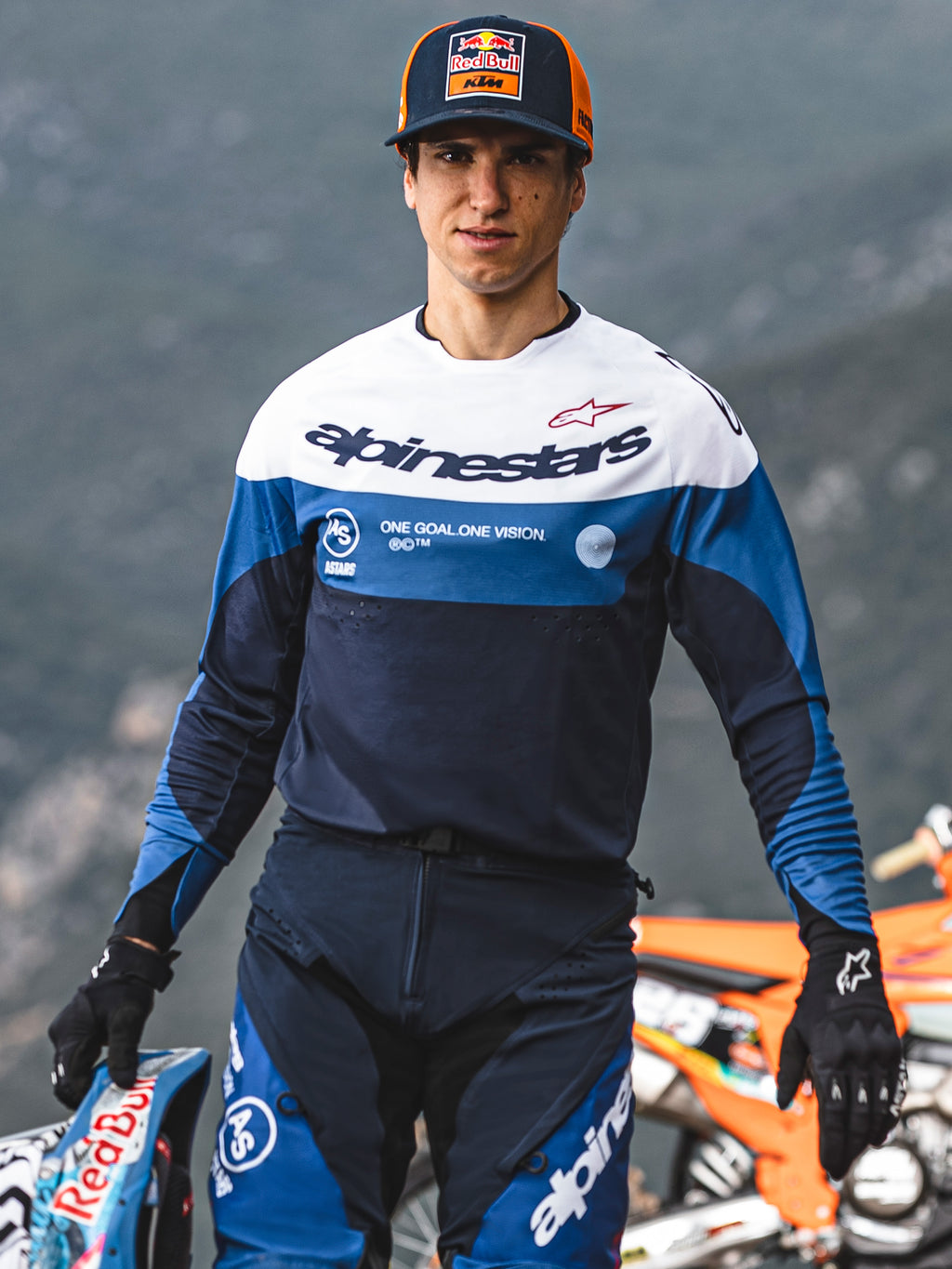 Motorcrosser in Alpinestars Pro-Dura Jersey en Broek in Navy Bright Blue en wit, voorzien van 'One Goal One Vision' branding, zwarte handschoenen, houdt een racehelm vast, professionele off-road race-uitrusting