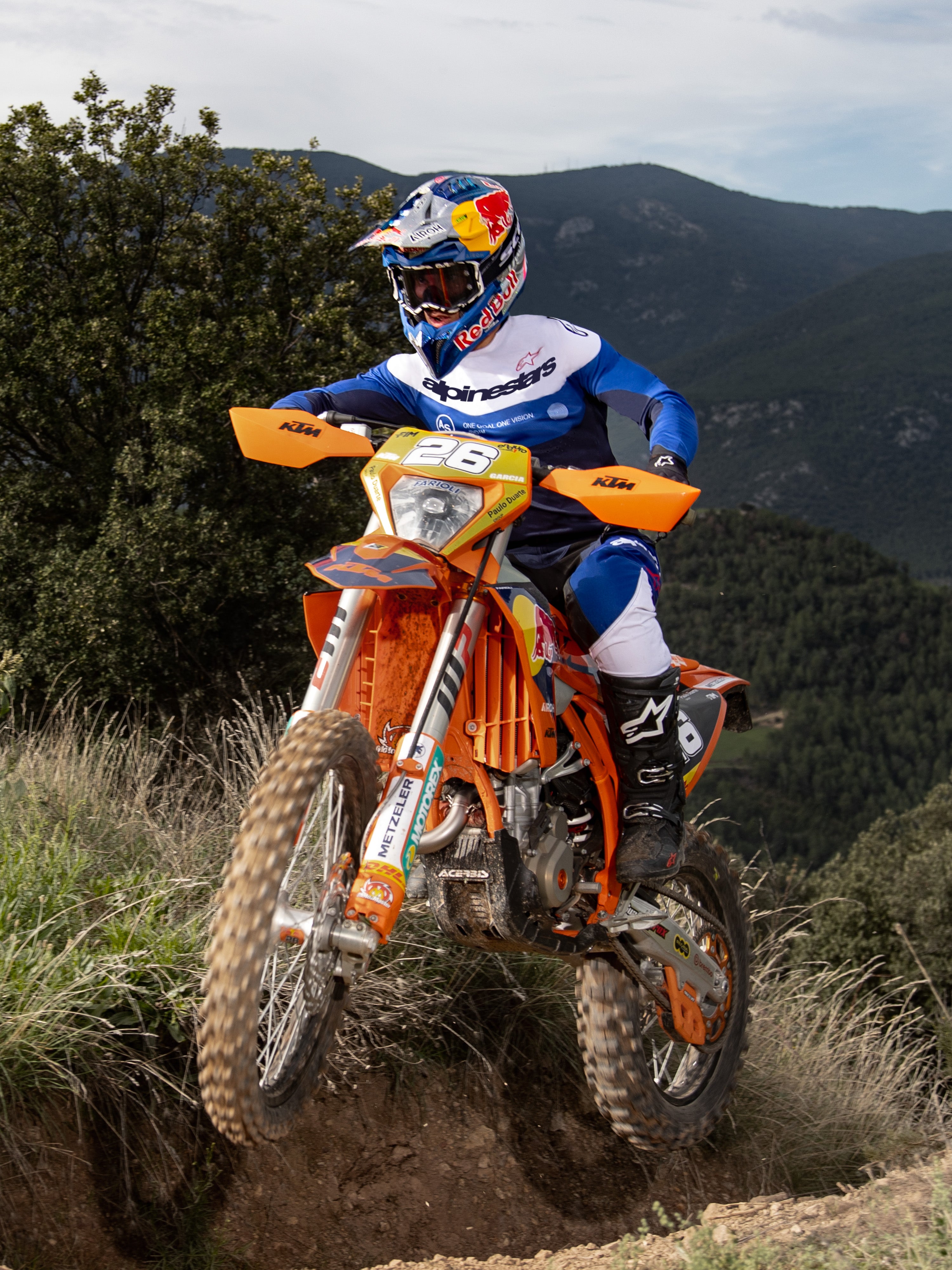 Enduro-rijder op een oranje crossmotor, Alpinestars Pro-Dura Jersey in Navy Bright Blue White, zwart-witte crossbroek en zwarte Alpinestars laarzen, rijdend op een ruig pad met bergen en bomen op de achtergrond.