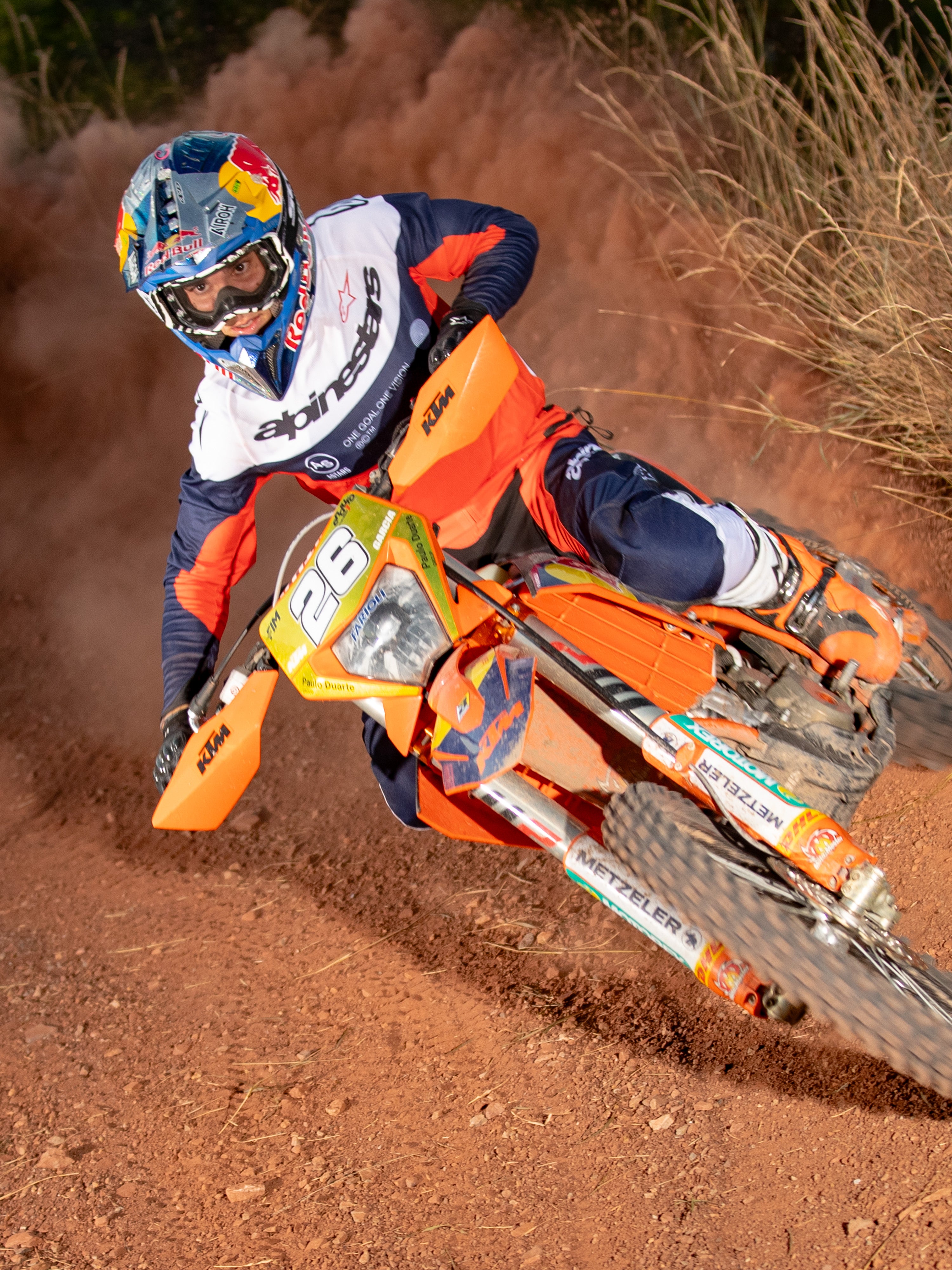 Motorcrosser die door een bocht leunt op een oranje motorfiets, gekleed in Alpinestars Pro-Dura jersey en broek in marineblauw, fel oranje en wit, bijpassende off-road race-uitrusting, Red Bull-helm, witte laarzen, actie-opname op een stoffige onverharde baan.